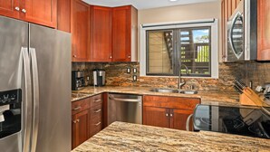 A kitchen with wooden cabinets, a stainless steel refrigerator, a dishwasher, a granite countertop, and a window with a view of greenery. - Summer Deal Oceanfront Kauai Condo with Heated Pool Beach Access (Lihue)