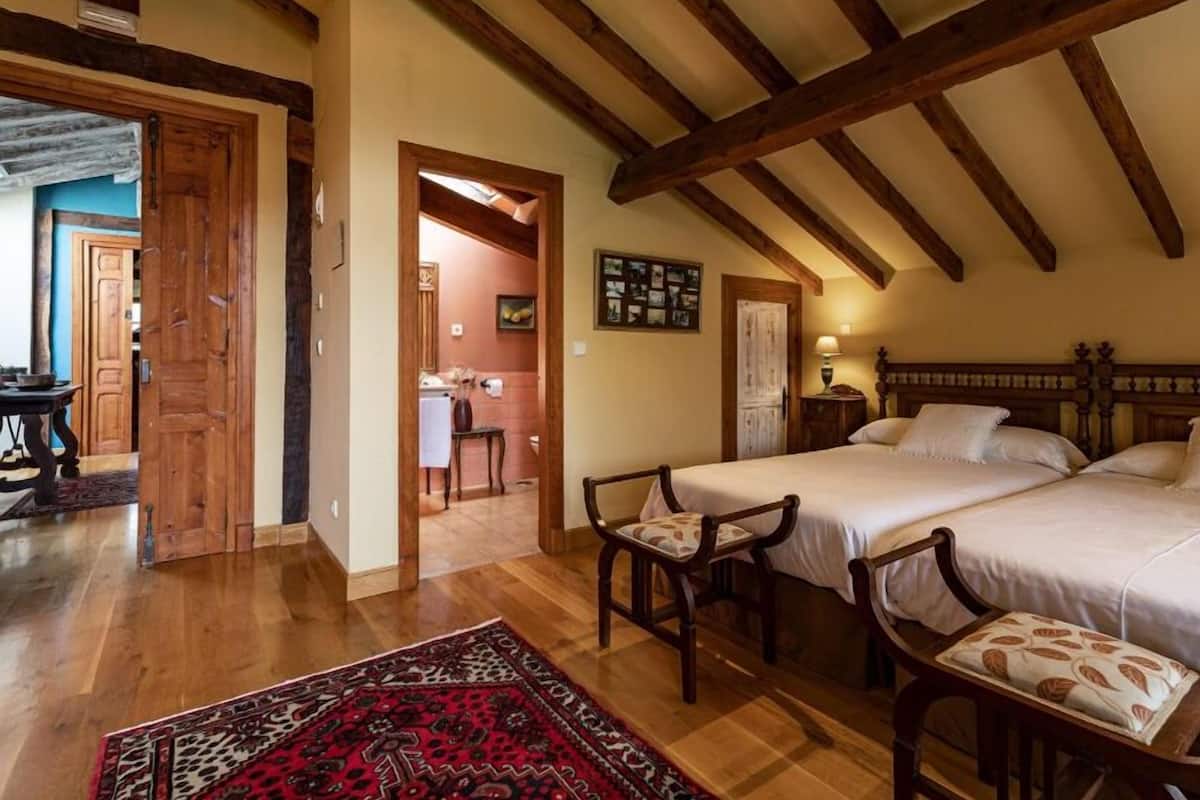 A bedroom with a wooden ceiling, a large bed, two chairs, a rug, and a bathroom visible through an open door.