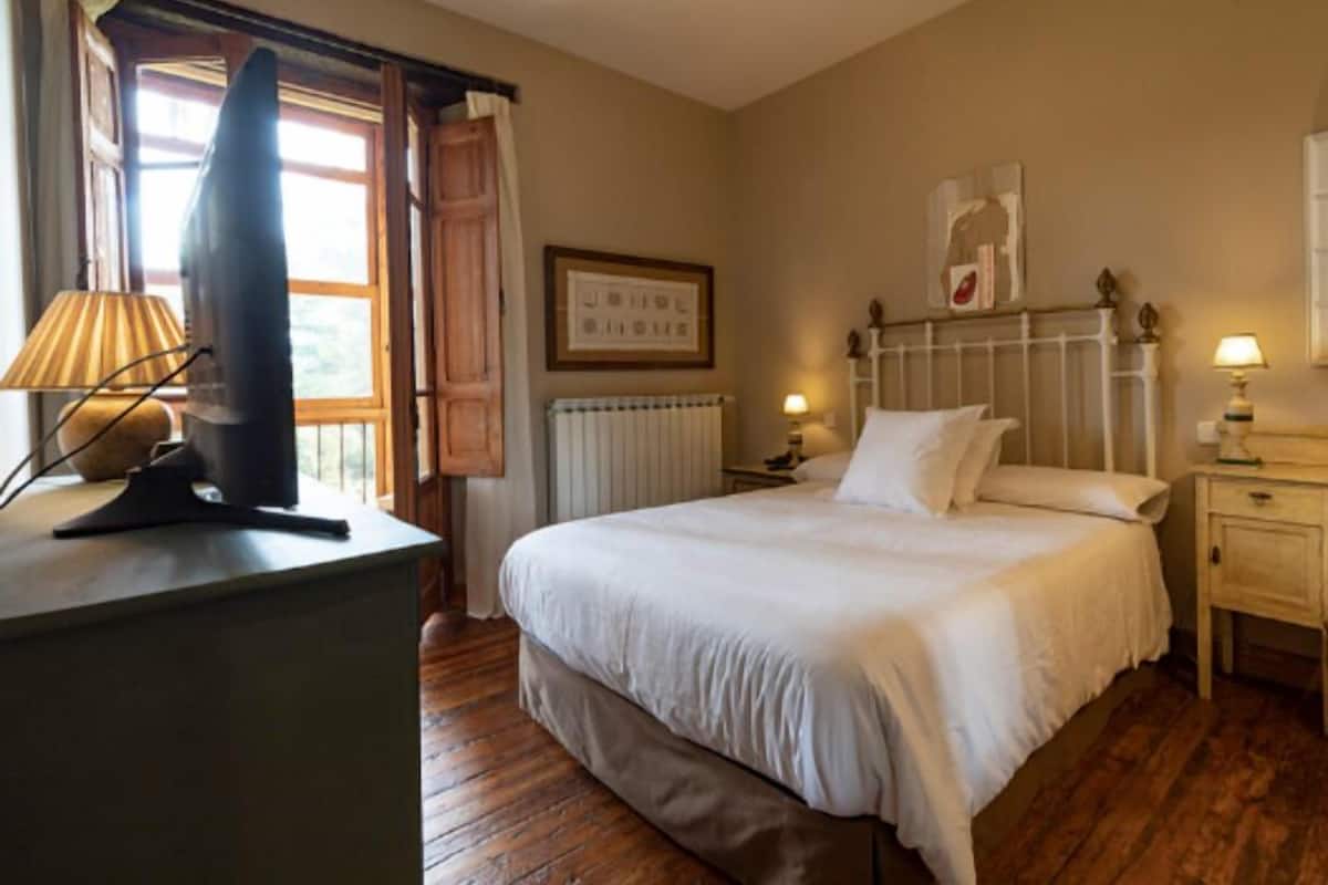 A bedroom with a bed, bedside tables, a lamp, a radiator, and a window with curtains.