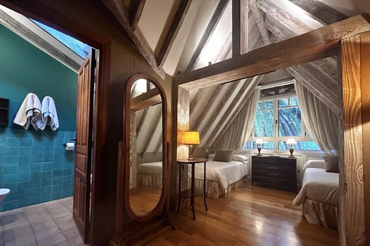 A bedroom with a large mirror, wooden beams, a window with curtains, and a bed with a nightstand.