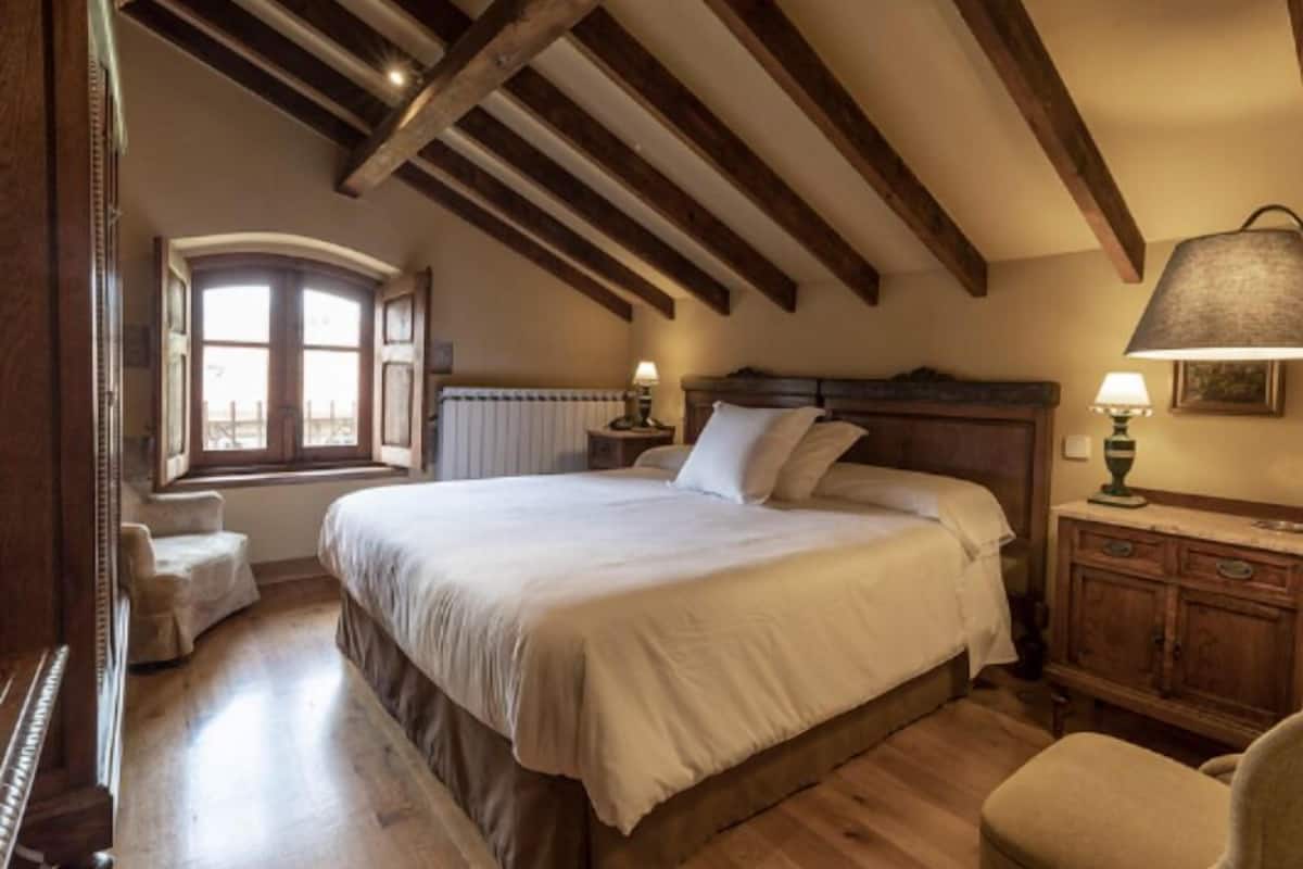 A bedroom with a large bed, wooden beams, a window, a radiator, and a nightstand with a lamp.