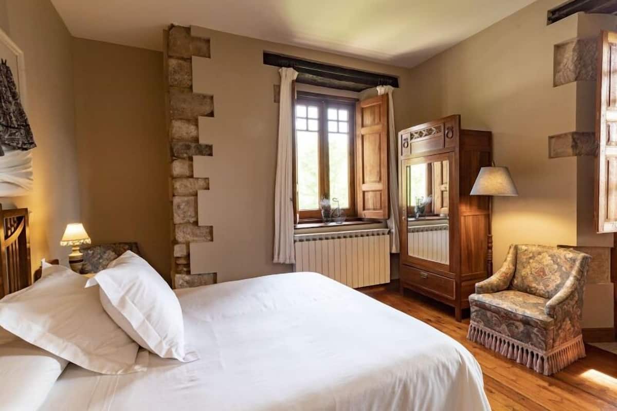 A bedroom with a large bed, a wooden chair, a radiator, and a window with curtains.