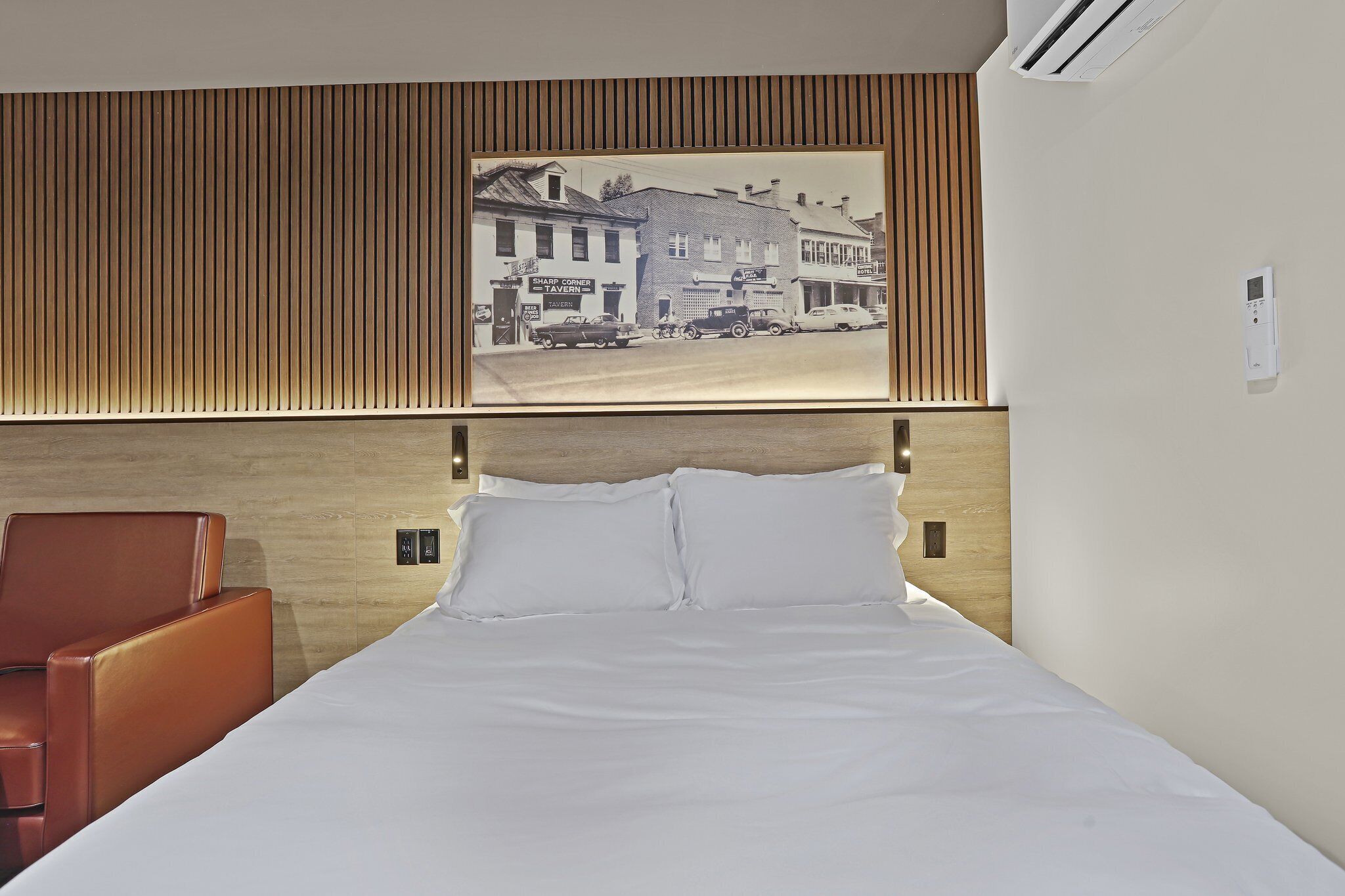 A hotel room with a bed, a brown leather chair, a wall-mounted air conditioner, and a framed picture of a street scene.