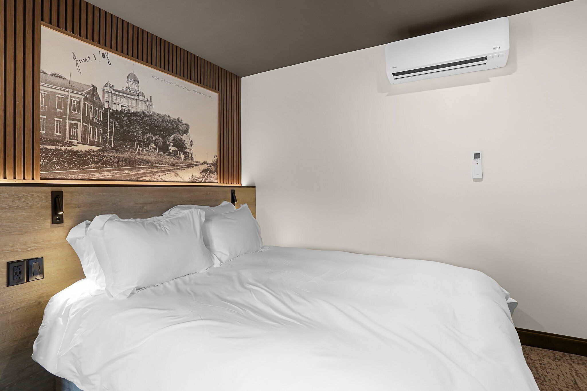A neatly made bed with white linens, a wall-mounted air conditioner, and a framed historical photograph above the bed.