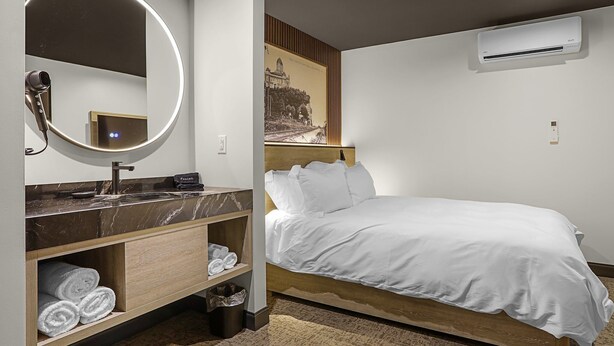 A modern hotel room with a marble bathroom vanity, a round mirror, a bed with white bedding, and a wall-mounted air conditioner. - The Hermann Motel (Hermann)