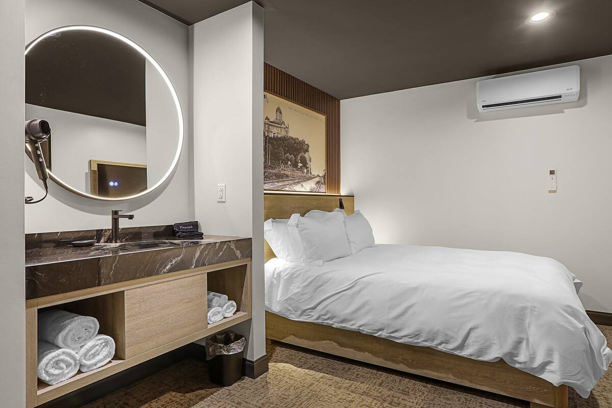 Une chambre d’hôtel moderne avec un meuble-lavabo en marbre, un miroir rond, un lit avec une literie blanche et un climatiseur fixé au mur.