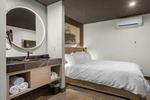 A modern hotel room with a marble bathroom vanity, a round mirror, a bed with white bedding, and a wall-mounted air conditioner. - The Hermann Motel (Hermann)