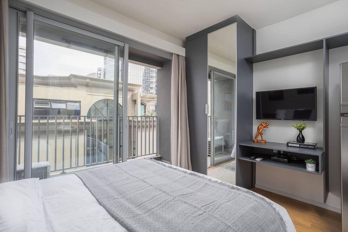 Habitación de hotel moderna con una cama grande, un televisor en estante empotrado y un balcón con vista a la ciudad.
