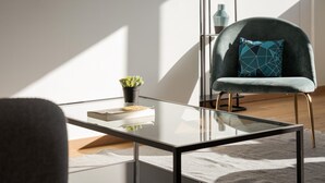 Ein modernes Wohnzimmer mit einem Glastisch, einem bequemen Sessel mit Kissen in geometrischem Muster und einem kleinen Blumentopf.