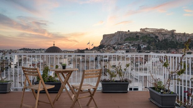 Eine Terrasse mit Holzsesseln und Tisch, mit Blick auf eine historische Stadtlandschaft mit antiken Ruinen.