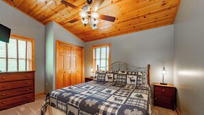 Une chambre avec un plafond en bois, un lit recouvert d’une courtepointe à motifs animaliers, une commode et une table de chevet avec une lampe.