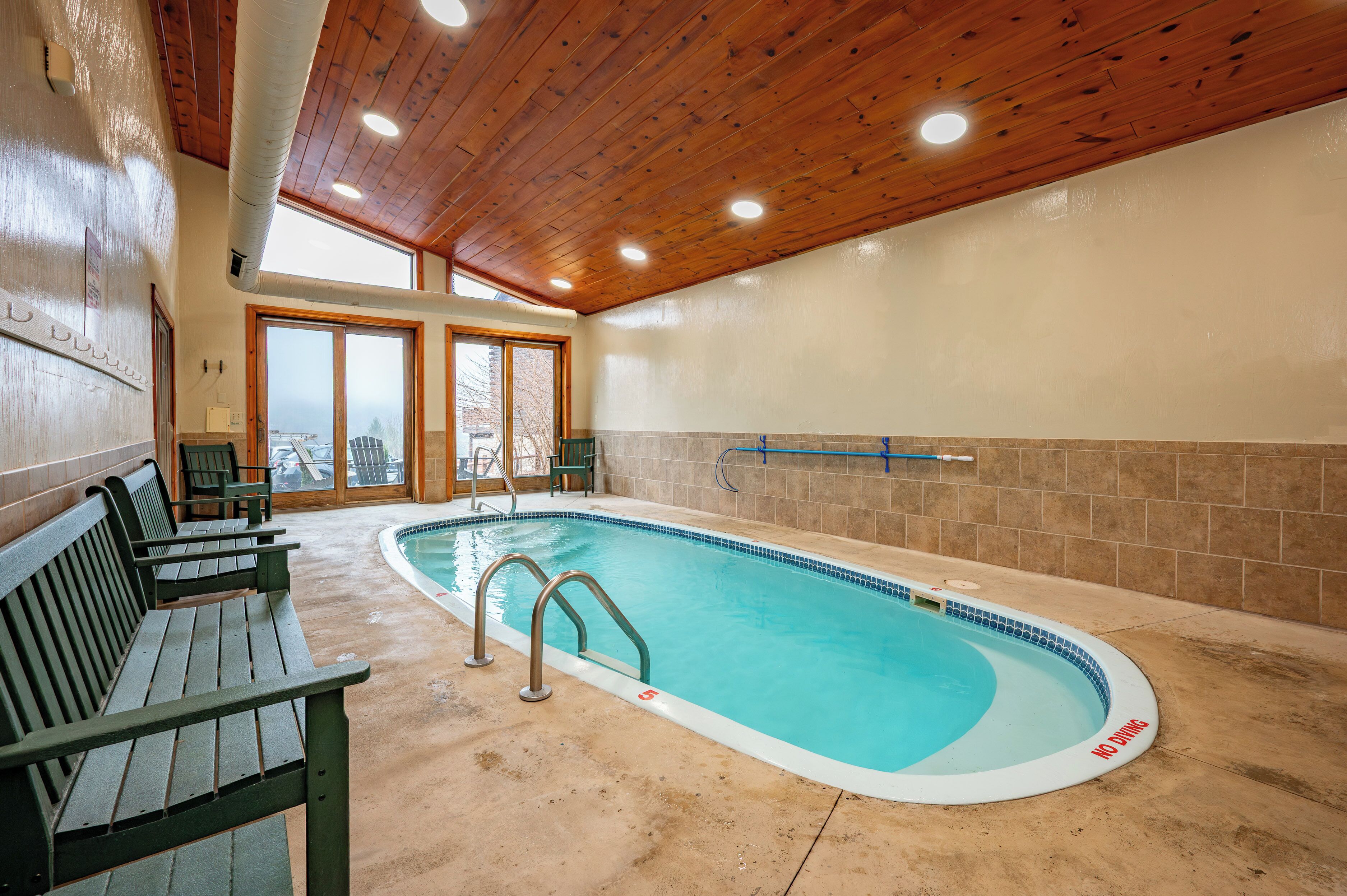 An indoor swimming pool with wooden benches, a tiled wall, and a view through large windows.