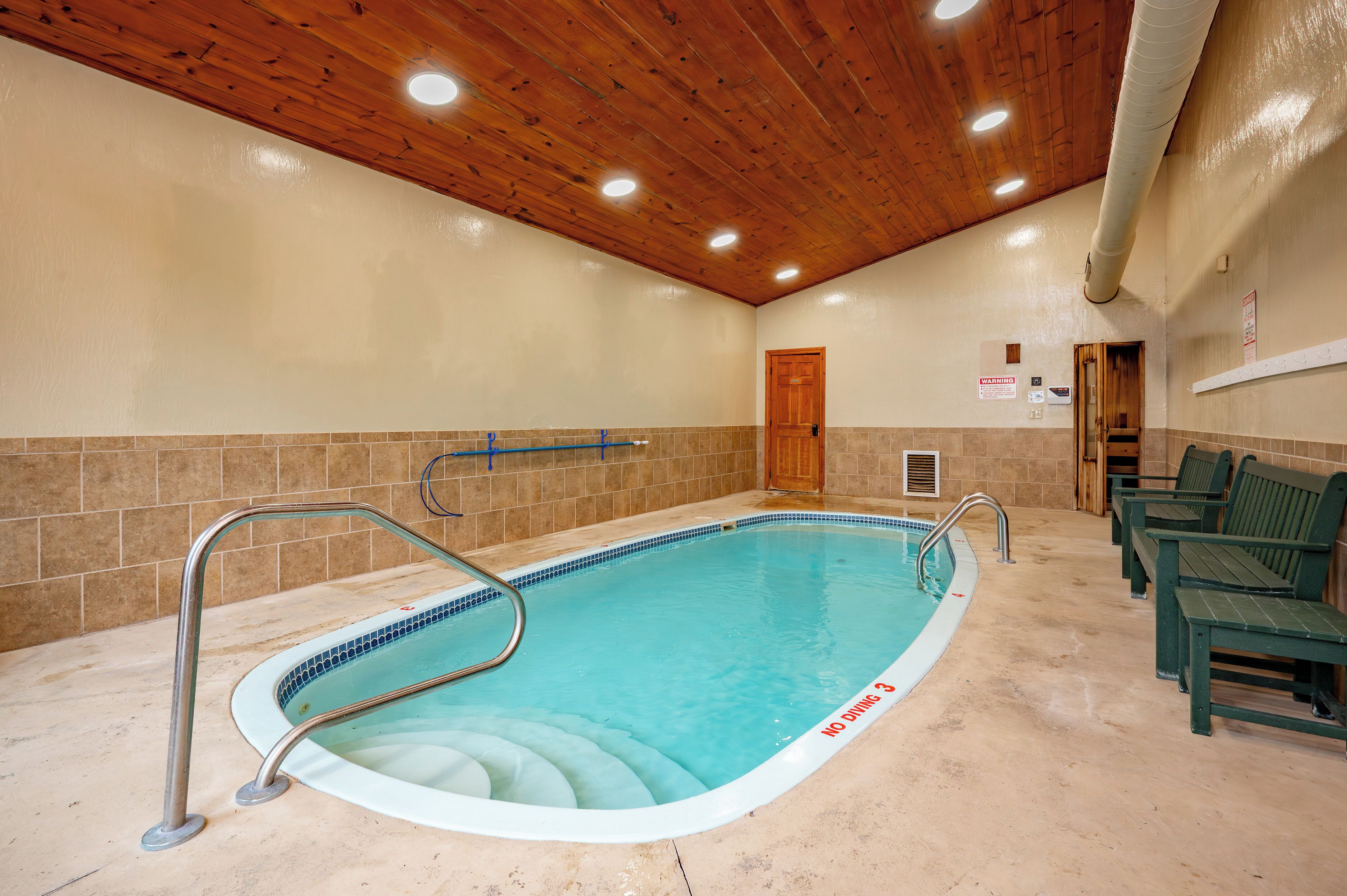 Ein Hallenbad mit Holzbalkendecke, gefliesten Wänden und einer Sitzgelegenheit am Beckenrand.