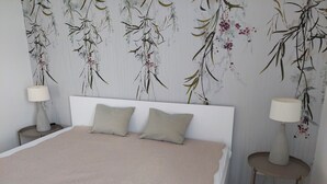 A bedroom with a bed, two pillows, a bedside table with a lamp, and a wall with a floral pattern. - 2-room vacation apartment directly on Lake Constance with bathing area (Überlingen)