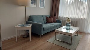 A modern living room with a blue sofa, a white coffee table, and a floor lamp. There are two framed pictures on the wall and a window with sheer curtains. - 2-room vacation apartment directly on Lake Constance with bathing area (Überlingen)