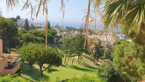 Üppige grüne Landschaft mit einem gepflegten Rasen, einem großen Baum und Blick auf eine Küstenstadt im Hintergrund.