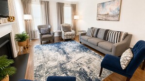 A living room with a fireplace, a ceiling fan, a sofa, two armchairs, a patterned rug, and a painting on the wall.