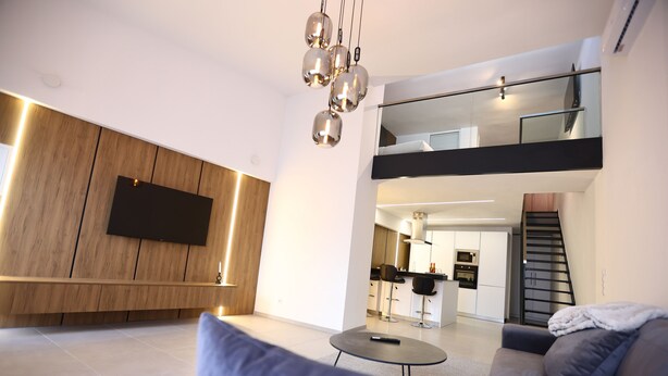 A modern living room with a sofa, a coffee table, and a TV mounted on a wooden wall. There is a staircase leading to an upper level with a balcony. - Moderne Loft Wohnung Koblenz (Koblenz)