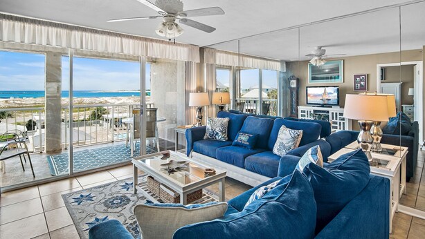 Een ruime woonkamer met een grote blauwe bank, een salontafel en uitzicht op het strand door grote ramen.