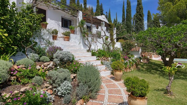 Una villa bianca con gradini in pietra che conducono a una porta in legno, immersa in un giardino curato con diverse piante e un cielo sereno.