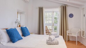 A hotel room with a bed, a chair, a window with curtains, and a wall-mounted air conditioner. - The Little Well: Private Pool, Sleeps2, Views (Frigiliana)