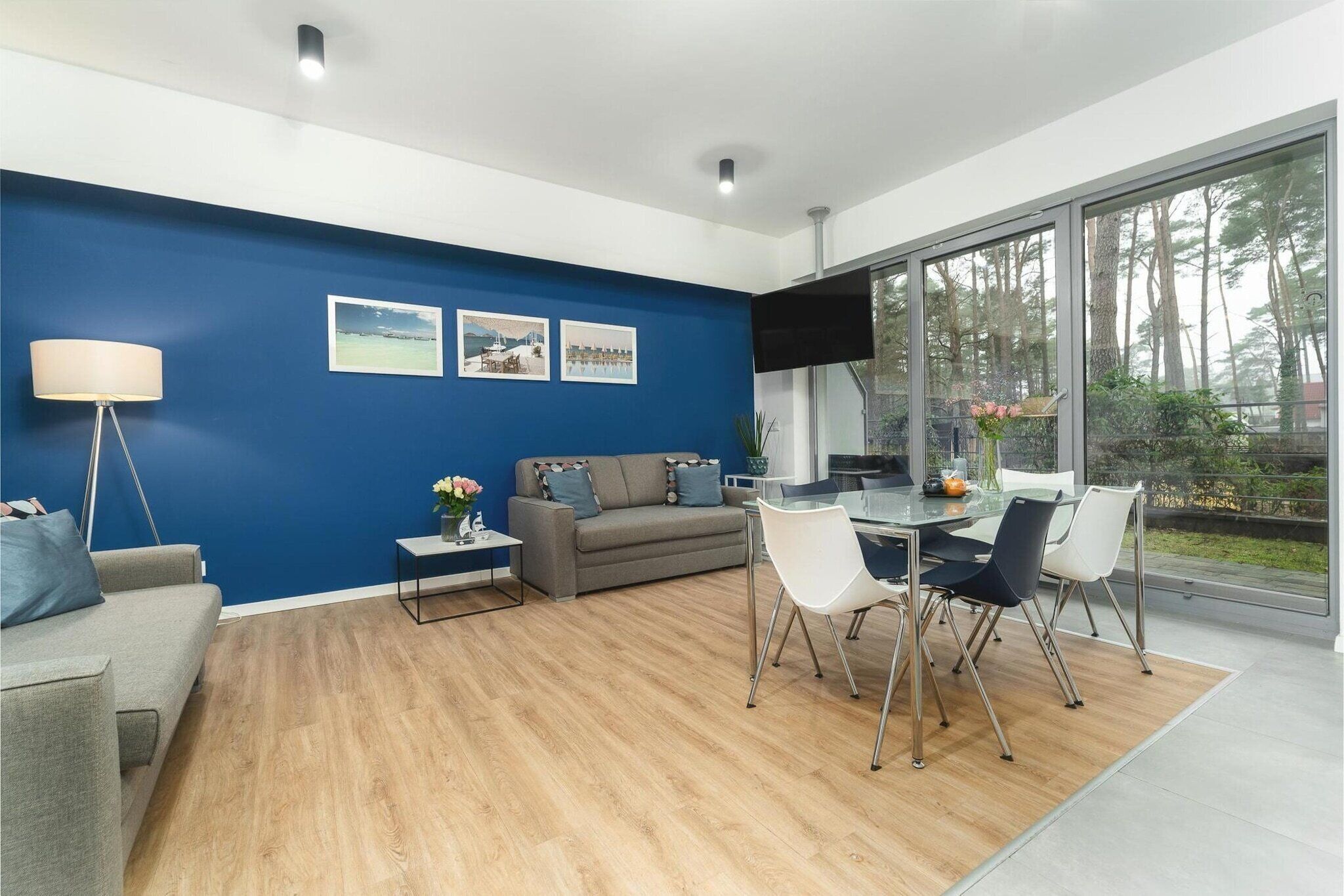 A modern living room with a blue accent wall, a grey sofa, a dining table with chairs, and a large window offering a view of trees.