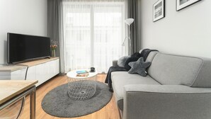 A modern living room with a grey sofa, a flat-screen TV, a coffee table, and a large window with sheer curtains. - Comfortable holiday apartment by the beach (Pobierowo)
