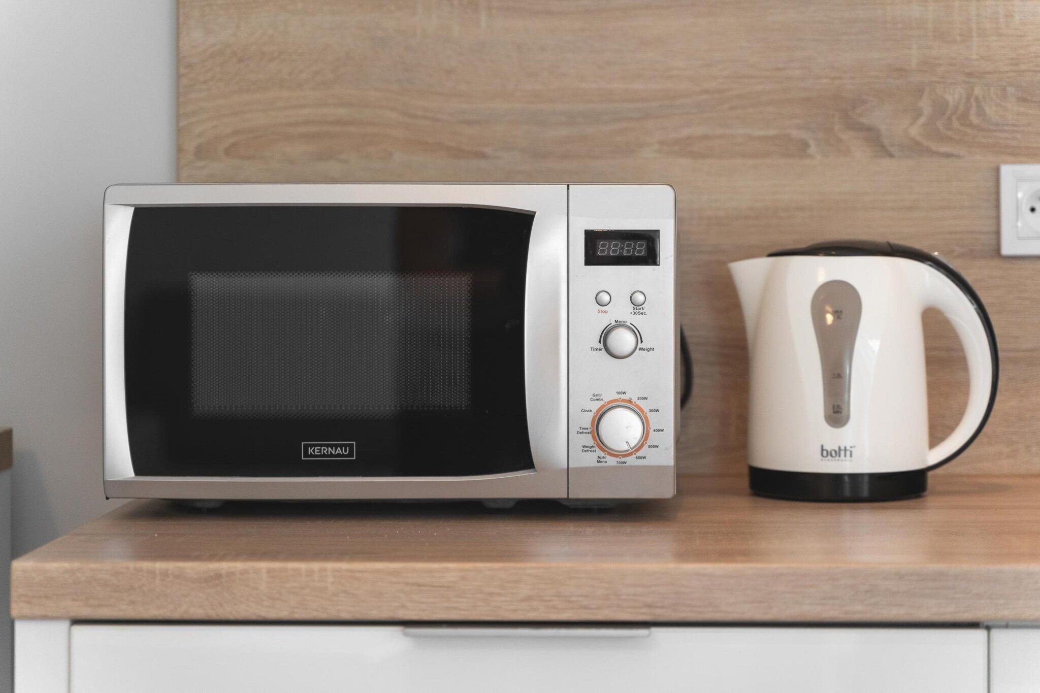 Un horno microondas con pantalla digital y panel de control, una tetera y una encimera de madera.