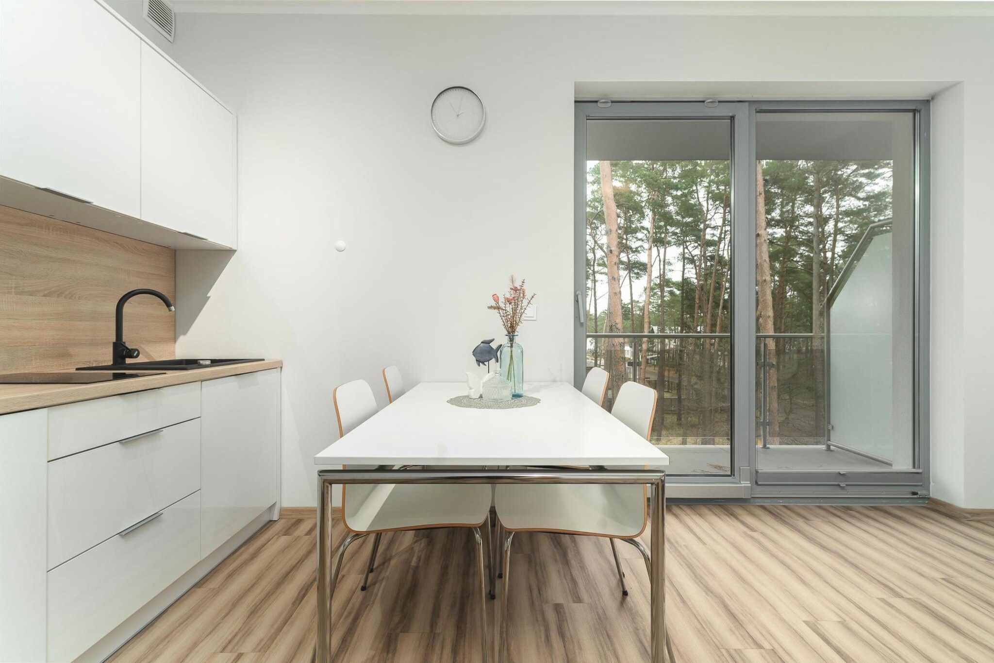 A modern kitchen with a dining table, chairs, and a view of trees through a glass door.