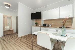 A modern kitchen with white cabinets, a flat-screen TV, a microwave, and a dining area with a white table and chairs. - Holiday studio very close to the seaside beach (Pobierowo)