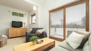 A modern living room with a sofa, a coffee table, a TV, and a window with blinds. - Apartment in Rewal by Baltic Sea Beach (Rewal)