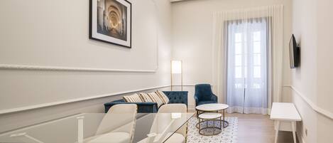 Una sala de estar moderna con una mesa de centro de vidrio, un sofá, un sillón azul y una pequeña mesa blanca.