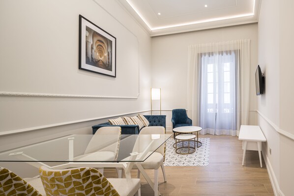 Una sala de estar moderna con una mesa de centro de vidrio, un sofá, un sillón azul y una pequeña mesa blanca.