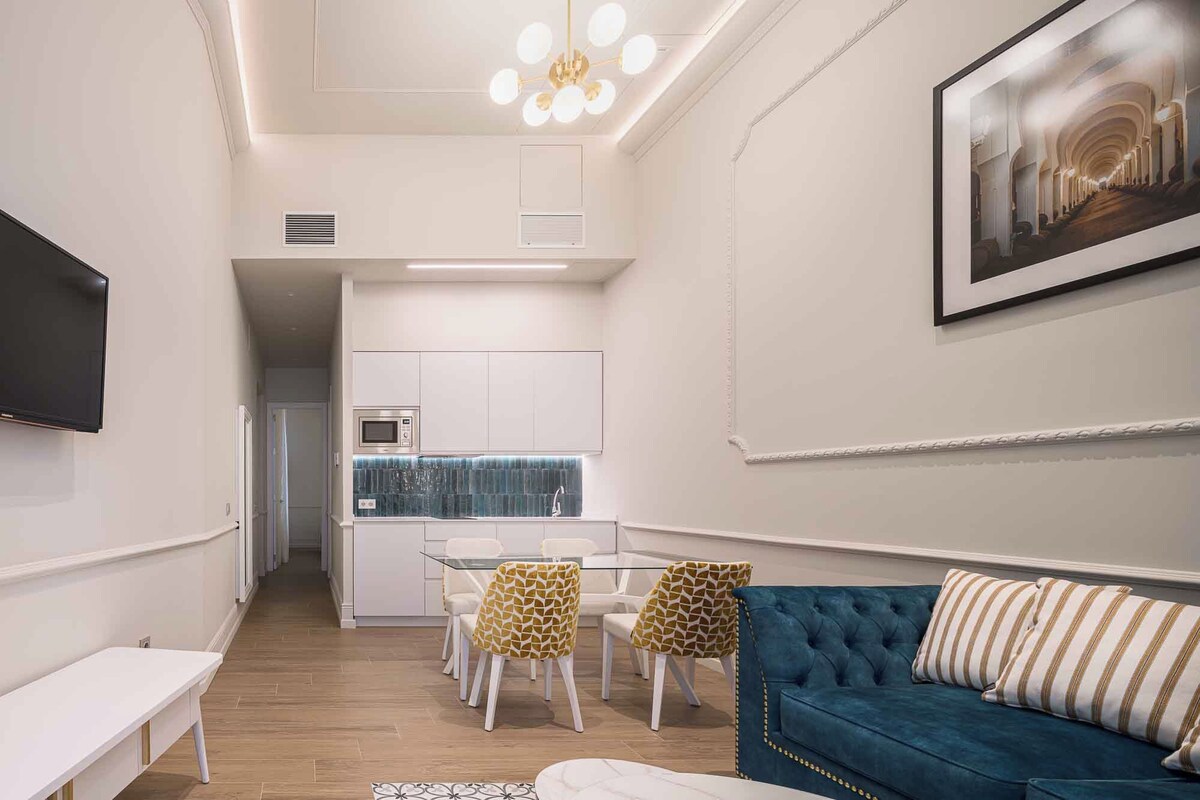 A modern living room with a blue sofa, a dining table with patterned chairs, a flat-screen TV, and a kitchen area with white cabinets and a blue backsplash.