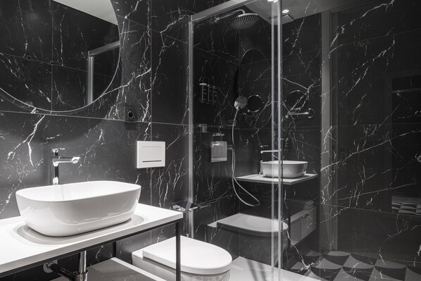 A modern bathroom with a white sink, toilet, and shower area, featuring marble walls and a glass-enclosed shower.