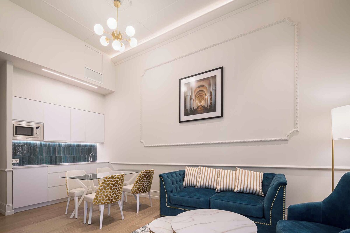 Uma sala de estar moderna com um sofá azul, uma mesa de jantar de vidro e uma área de cozinha com armários brancos e um micro-ondas.