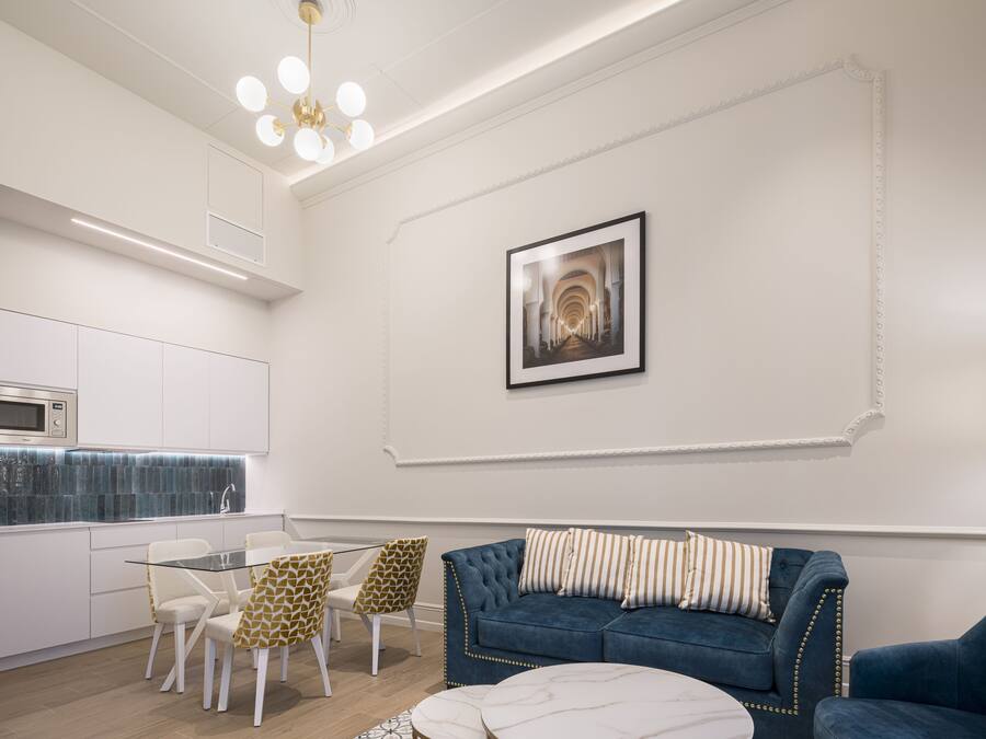 A modern living room with a blue sofa, white chairs, a glass coffee table, and a kitchen area with white cabinets and a blue backsplash.
