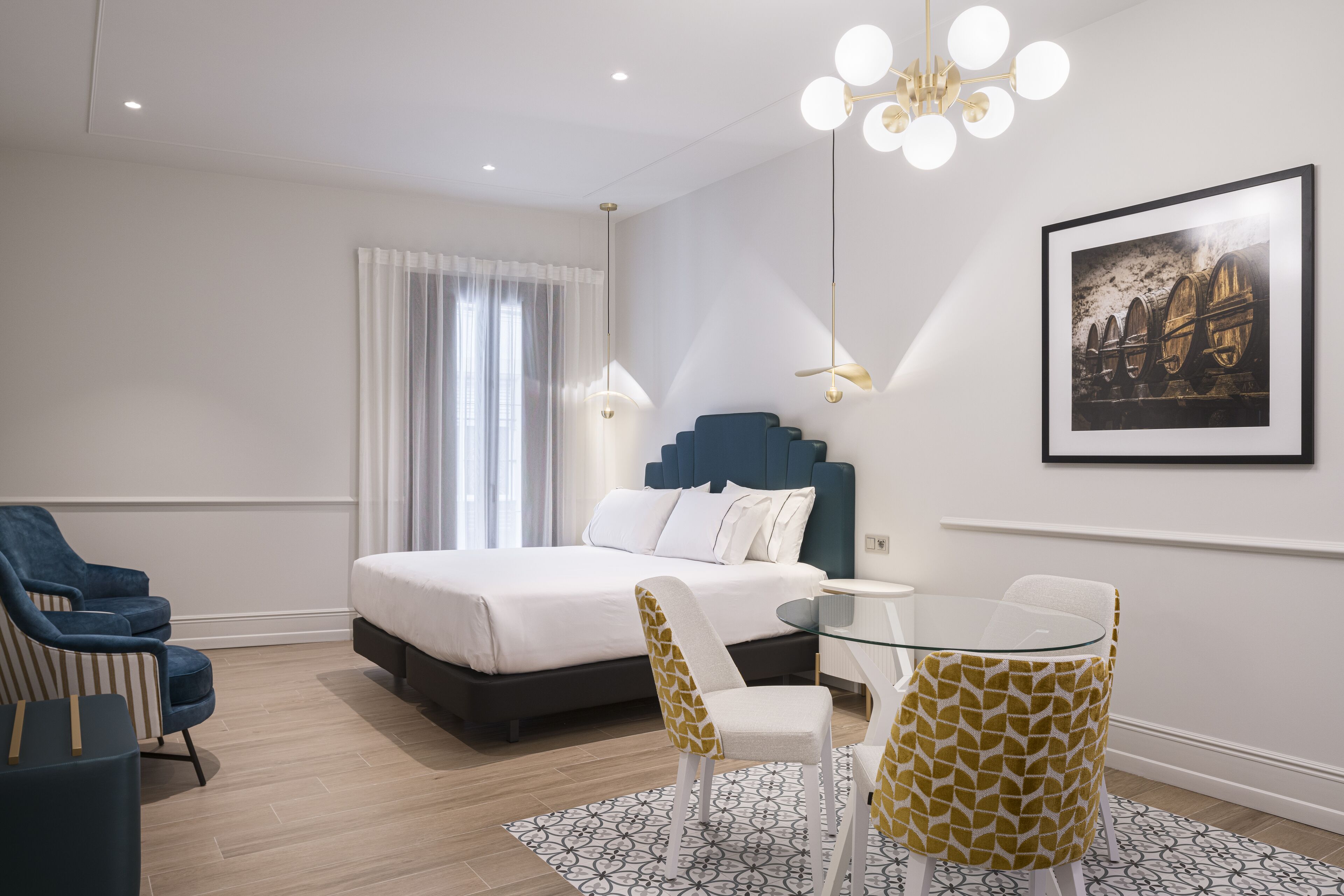 A modern hotel room with a large bed, a glass table with chairs, a blue armchair, and a chandelier.