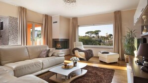 A modern living room with a sofa, armchair, and ottoman, a fireplace, and a large window with a view of trees and buildings. - Charming Villa Diora with a swimming pool and wellness area (Karojba)