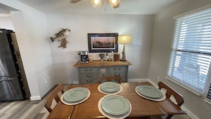 Un coin repas avec une table en bois dressée pour quatre personnes, un ventilateur de plafond et une fenêtre avec des stores.