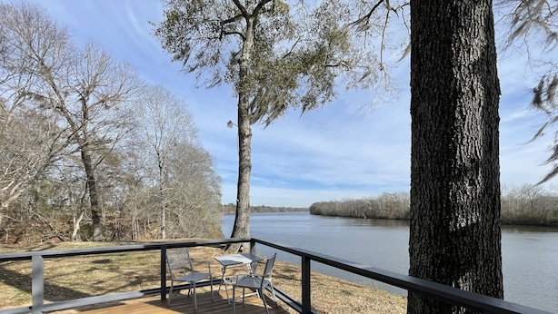 Une terrasse en bois avec des rampes en métal, une table et des chaises, donnant sur une rivière paisible bordée d’arbres.