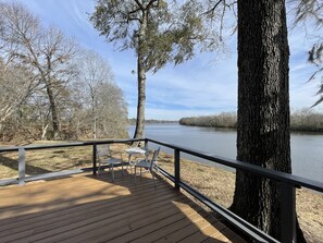 Une terrasse en bois avec des rampes en métal, une table et des chaises, donnant sur une rivière paisible bordée d  ’arbres.