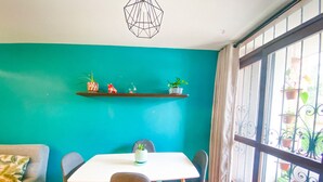 Un coin repas avec une table blanche et quatre chaises, un mur de couleur turquoise et une fenêtre avec des barreaux en métal.