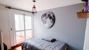 Une chambre à coucher avec un lit, une décoration murale représentant la lune, un luminaire suspendu et une étagère en bois sur laquelle est posée une plante.