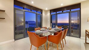 A dining area with a glass table and orange chairs, large windows with a city view, and a balcony with a railing.