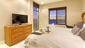 A bedroom with a TV on a wooden dresser, a bed with white bedding, and a window with a view.
