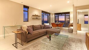 A modern living room with a glass coffee table, a brown sectional sofa, and a dining area with a view of the sunset.