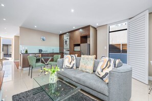 Ein modernes Wohnzimmer mit einem grauen Sofa, einem Glastisch und einer Küche im Hintergrund.