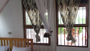 A room with two windows, each with a curtain featuring a leaf pattern. There is a wooden bed with white pillows and a small bedside table. - Nutmeg House Zanzibar (Jambiani)