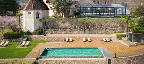 A pool area with lounge chairs, a stone wall, and a building with a glass greenhouse in the background. - Schloss Freudenstein (Appiano Sulla Strada del Vino)